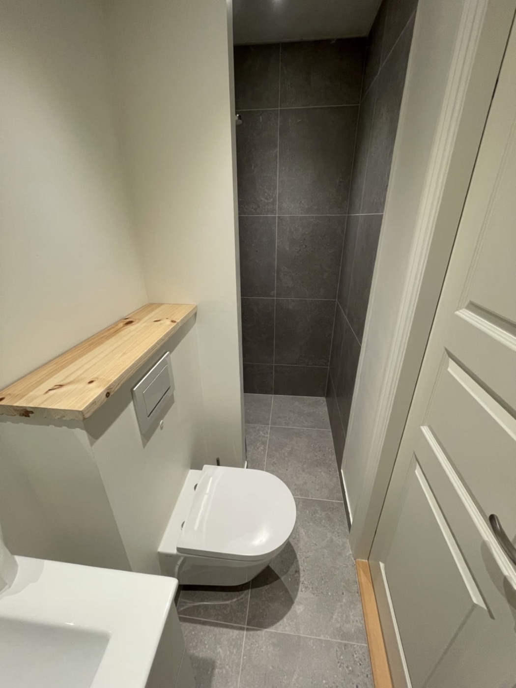 Gæstetoilet med vægophængt toilet, træhylde og mørkegrå fliser i brusenichen